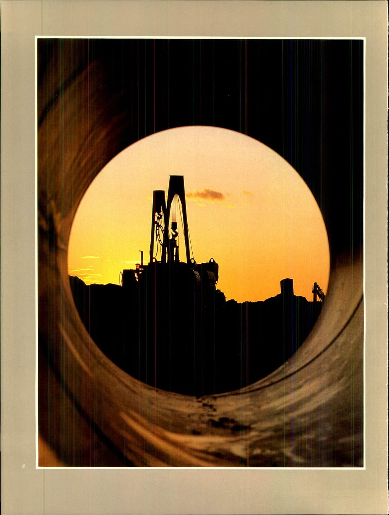 A pipelaying boom and derrick silhouetted through the mouth of a large-diameter pipe at sunset.
