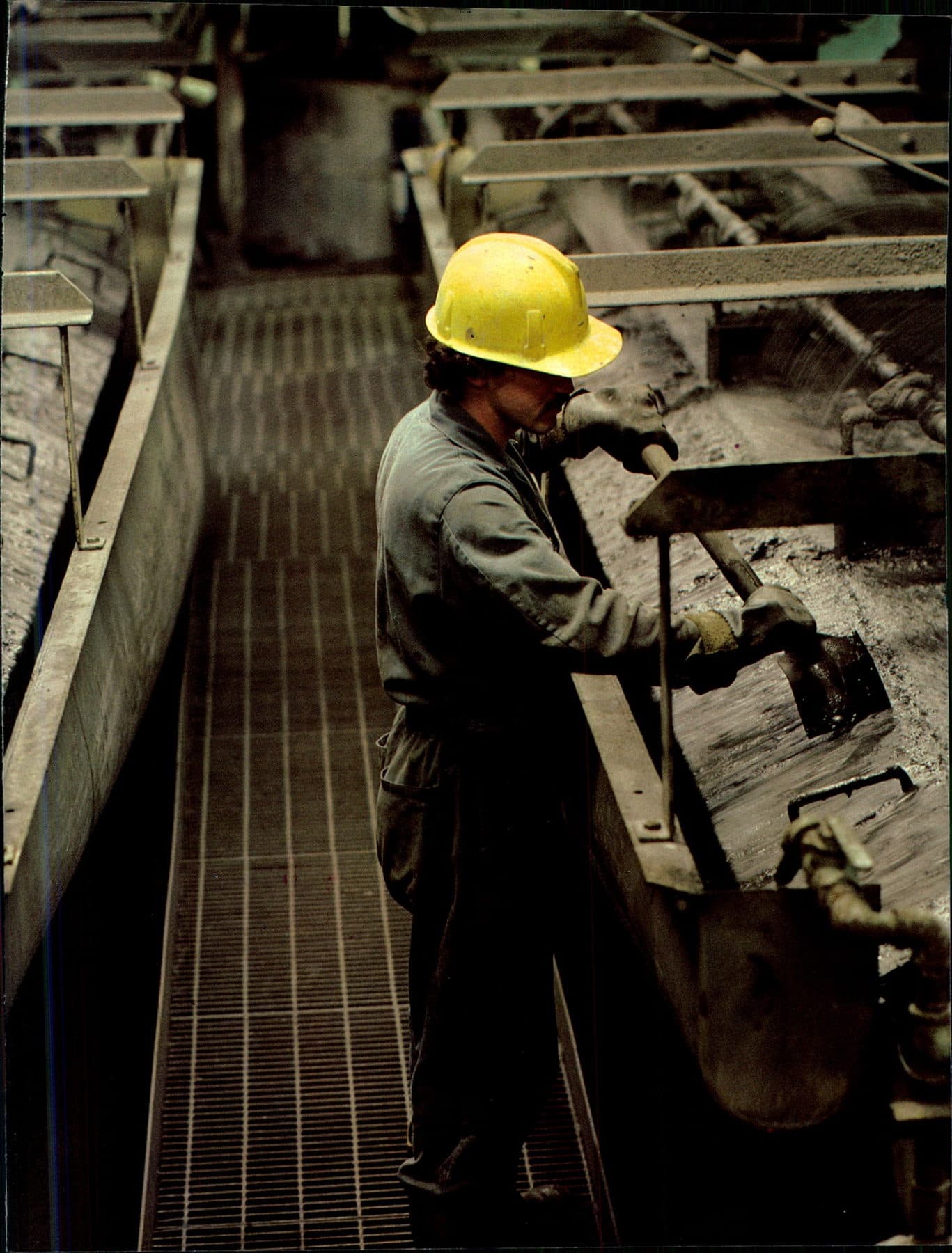 A mill worker in a yellow hard hat scrapes material in a long processing trough.
