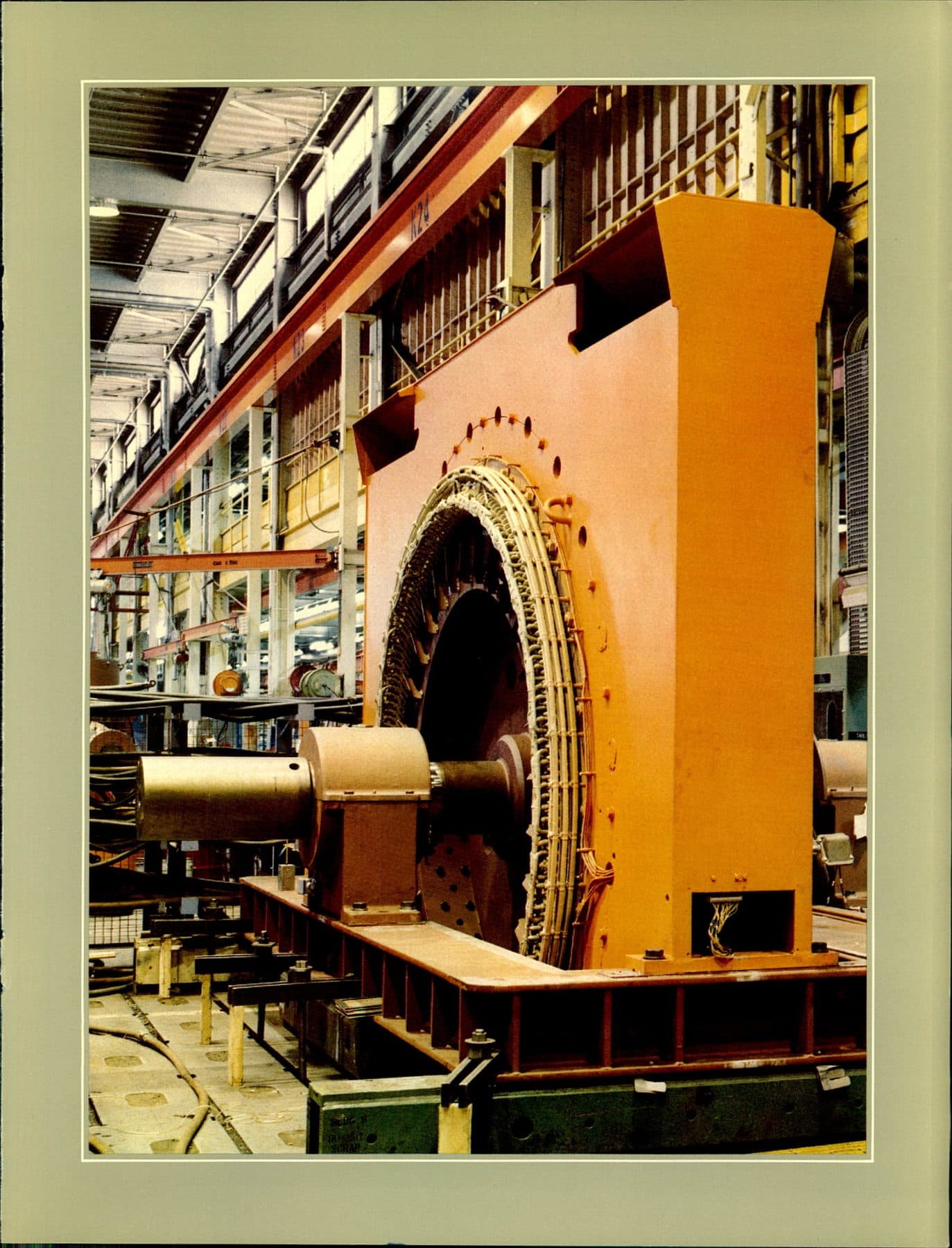A massive orange stator on a red steel bed, copper windings visible in the bore, factory bay behind.